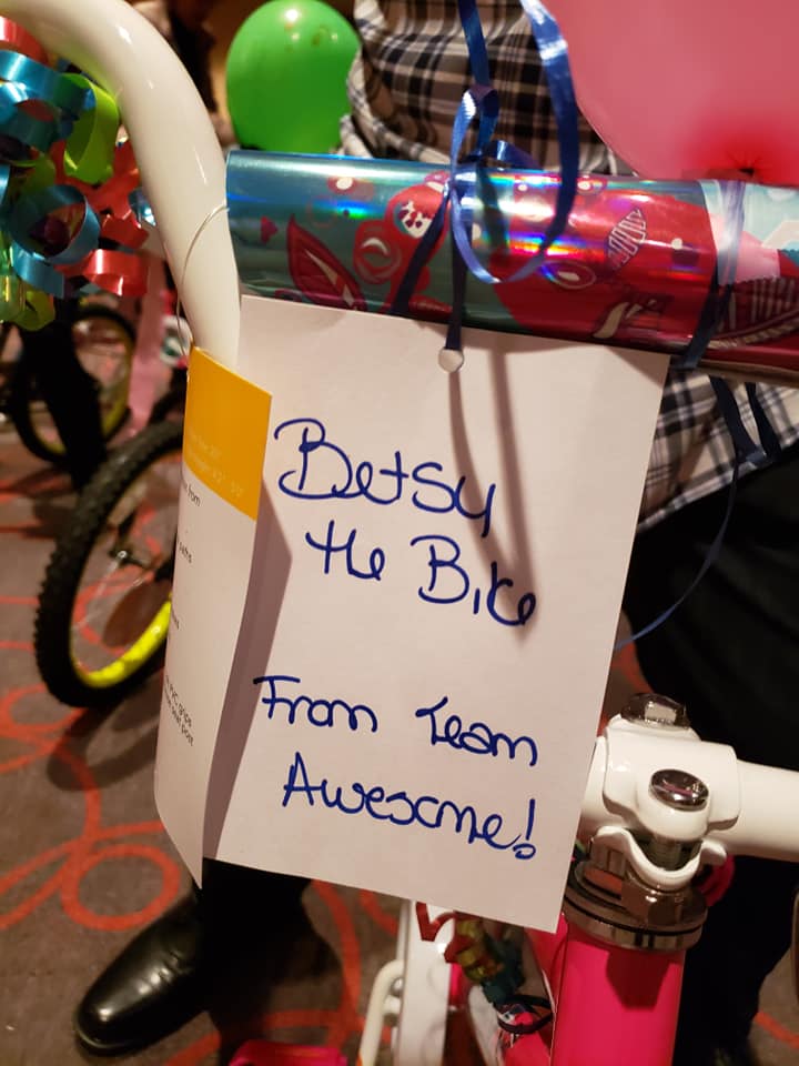 Close-up of a decorated bike handle with a handwritten note attached. The note reads 'Betsy the Bike, From Team Awesome!' The bike is wrapped in colorful paper, showing the completion of the assembly during a Charity Bike Build event.