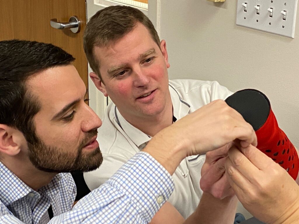 Two men collaborate on assembling a prosthetic hand. Both are focused and working closely together, one man holds a part of the prosthetic while the other assists in making adjustments. The atmosphere is one of teamwork and precision as they carefully work to ensure the hand is correctly assembled. This activity is part of a team building exercise aimed at making prosthetics for those in need.