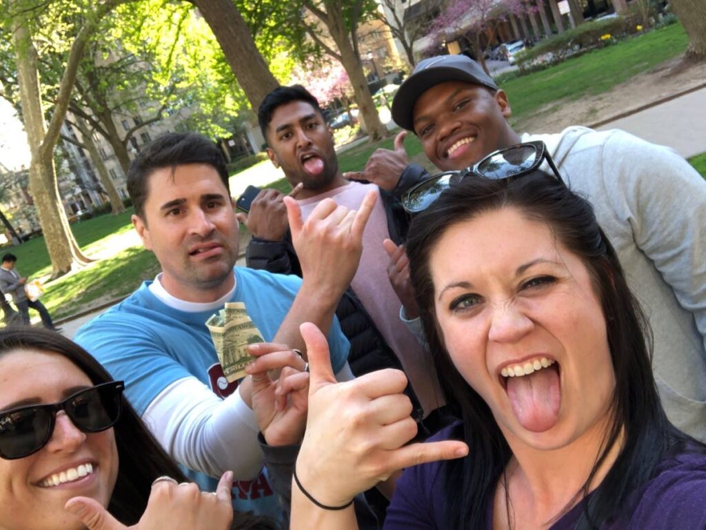 Team making funny faces and gestures, holding Monopoly money during a playful moment in the Team-opoly scavenger hunt event outdoors.