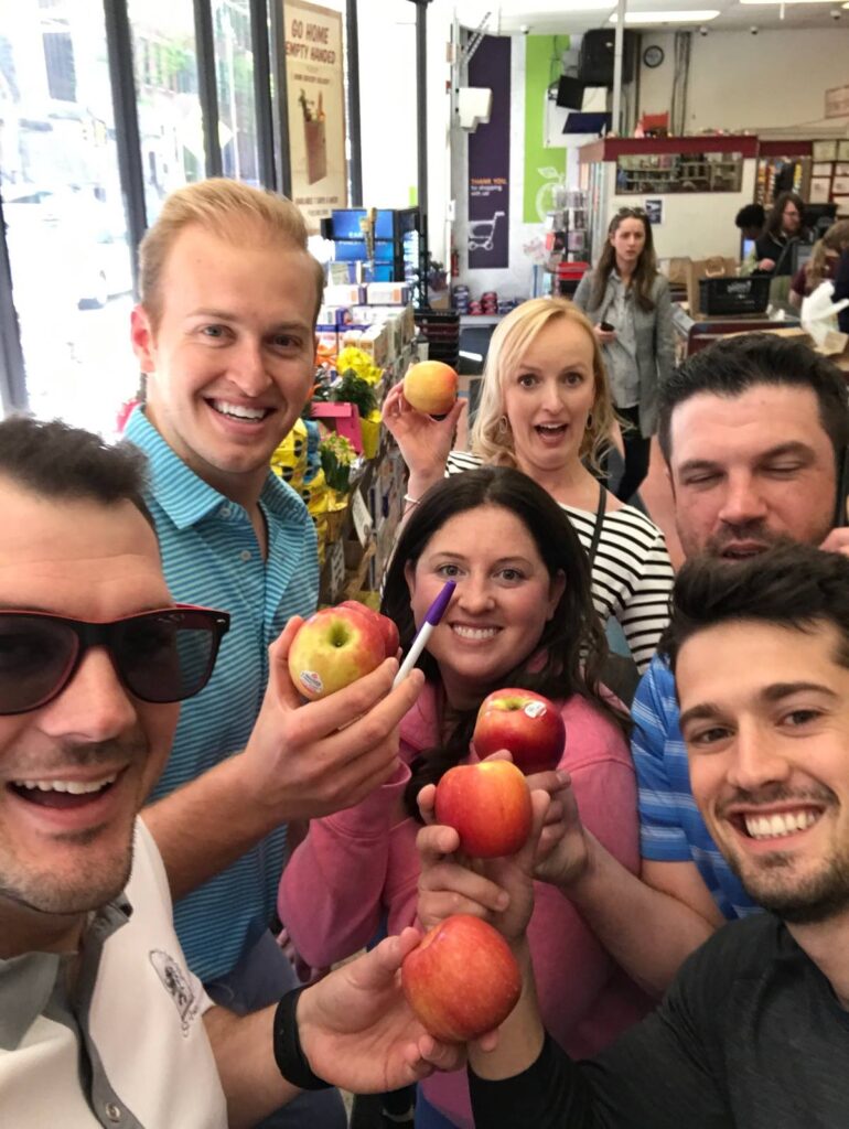 Team posing with apples inside a grocery store during a challenge for the Team-opoly scavenger hunt event.