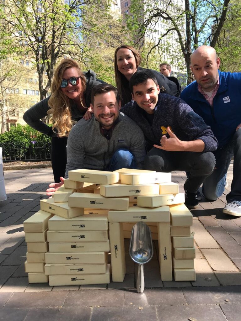 Team proudly posing behind a structure made from oversized wooden blocks during a challenge in the Team-opoly scavenger hunt event.