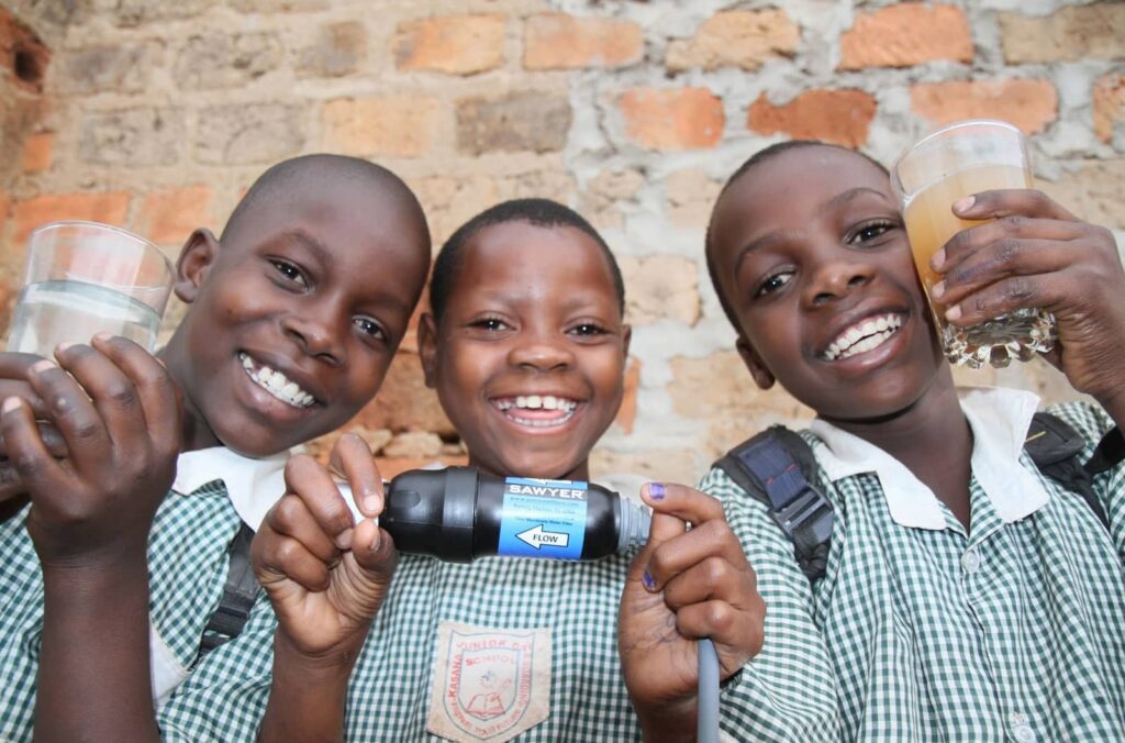 Three smiling children hold clean water glasses, showcasing the impact of Clean Water Connection's filtration systems. These systems provide access to safe drinking water in communities worldwide, helping empower people through clean water initiatives.