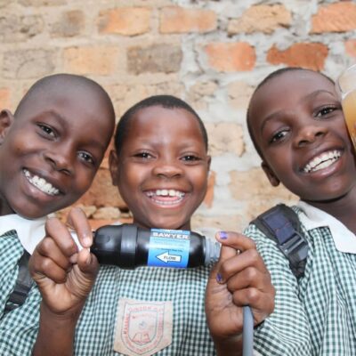 Three smiling children hold clean water glasses, showcasing the impact of Clean Water Connection's filtration systems. These systems provide access to safe drinking water in communities worldwide, helping empower people through clean water initiatives.