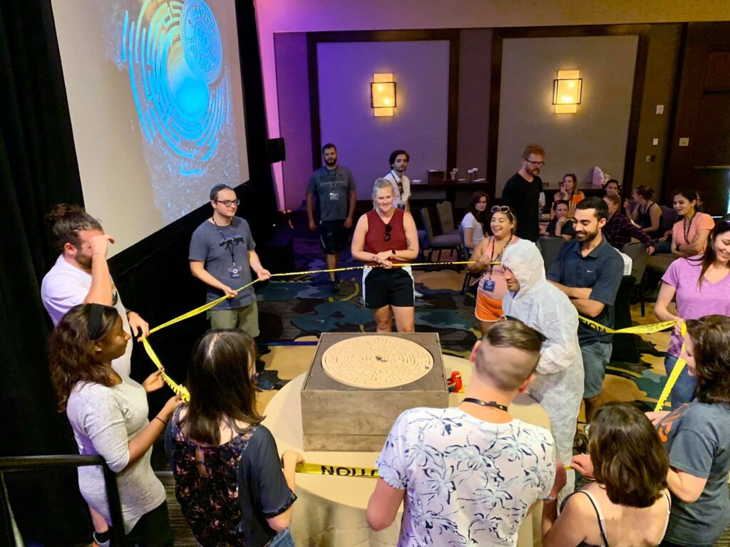 Group of participants gathers around a large maze puzzle table, focusing on solving the challenge together during the 'Escape The Maze' team building event, surrounded by caution tape for an immersive experience.