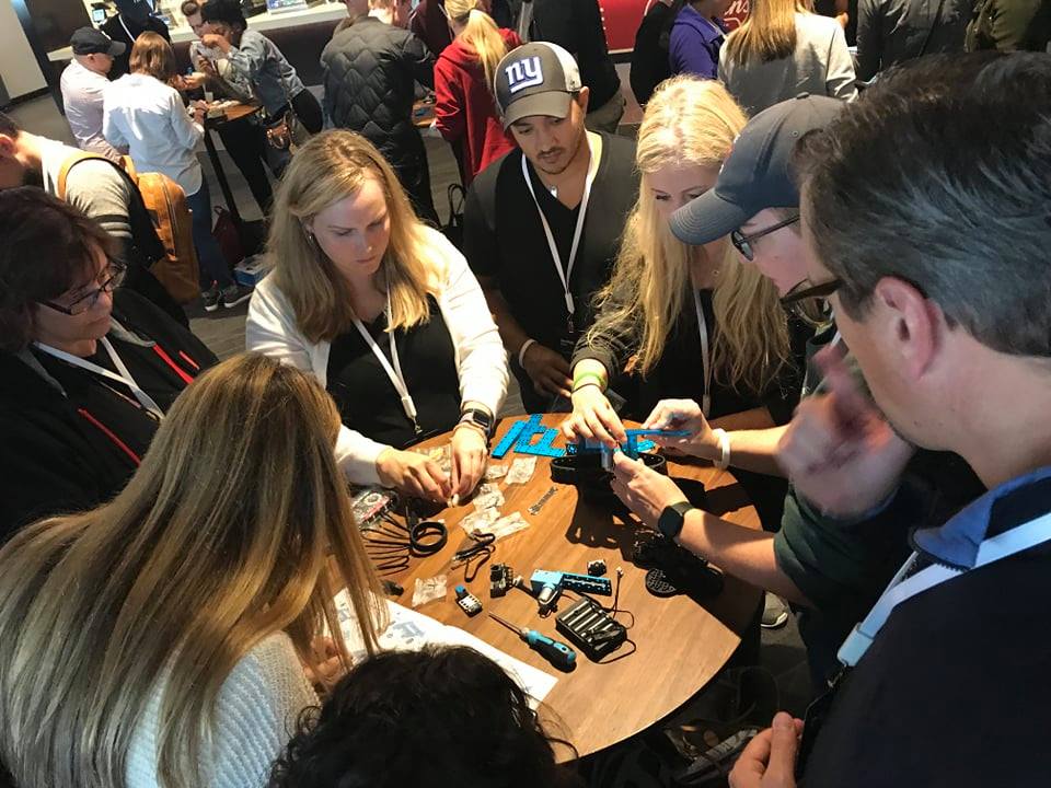 A group of participants is gathered around a table, working together to assemble robot parts. They are focused and engaged in a team building activity, using various tools and components to construct a functioning robot.