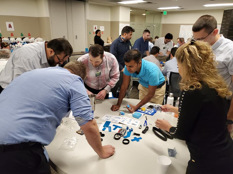 A group of participants is gathered around a table, fully engaged in assembling small robotic parts. The table is scattered with various components such as wheels, gears, and blue plastic pieces. Each person is focused on a task, contributing to the team effort to build a functioning robot during this team building event.