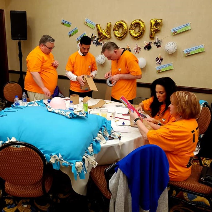 Group of participants in orange shirts working together to create pet care items during the Foster Pet Care Kits team building activity, with a large blue pet bed on the table and 'WOOF' balloons in the background.