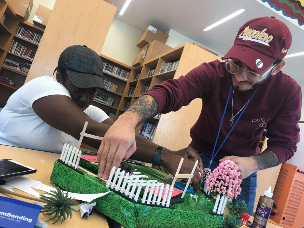 Two participants collaborate to build a detailed section of a miniature city during a City Build team building activity, adding fences and trees to the green landscape.