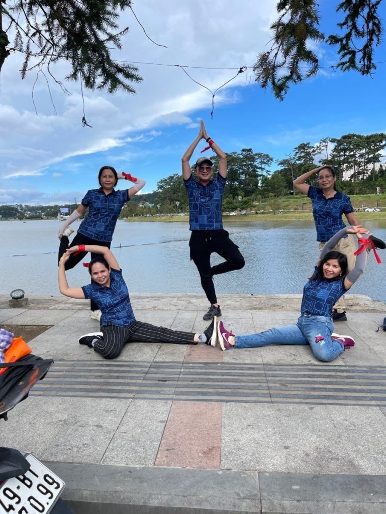 A team of five participants posing creatively during The Amazing Race Scavenger Hunt team building event, forming shapes with their bodies while outdoors near a lake.