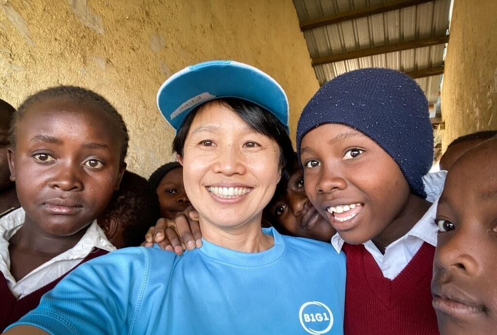 A woman in a blue B1G1 shirt and cap takes a selfie with a group of school children in red uniforms, who gather around her with bright smiles. This image reflects the positive impact of the donations on the local community.