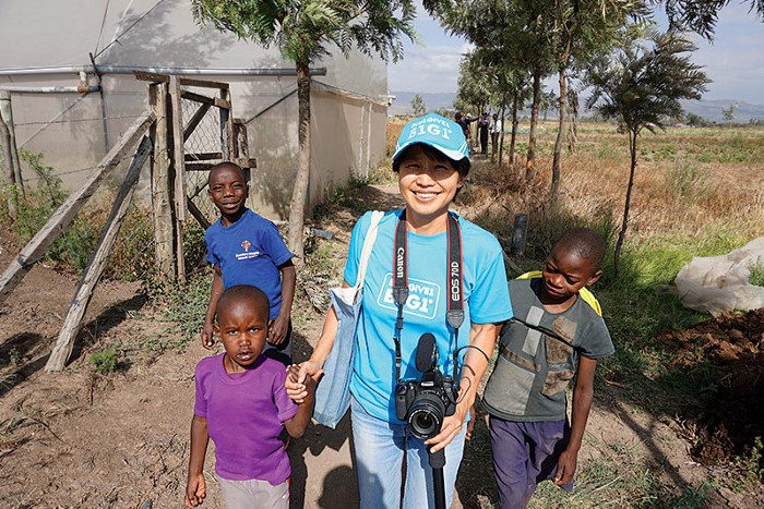 A woman wearing a blue B1G1 cap smiles warmly as she stands outside with three young children in a rural area. She is holding a camera, and the children, two boys and one girl, appear happy and curious. This photo captures a moment of connection and joy as donations are being delivered to support the community.