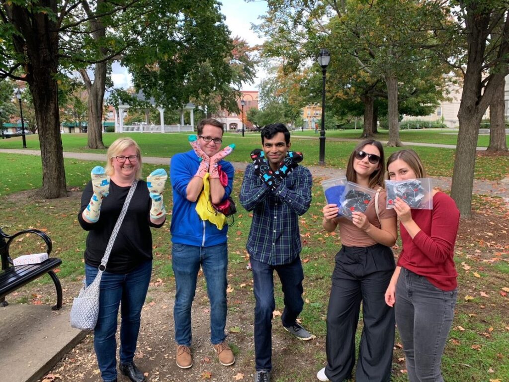 A group of five participants is standing outside in a park, each holding up socks and care items collected for a Healthcare Heroes scavenger hunt team building challenge.