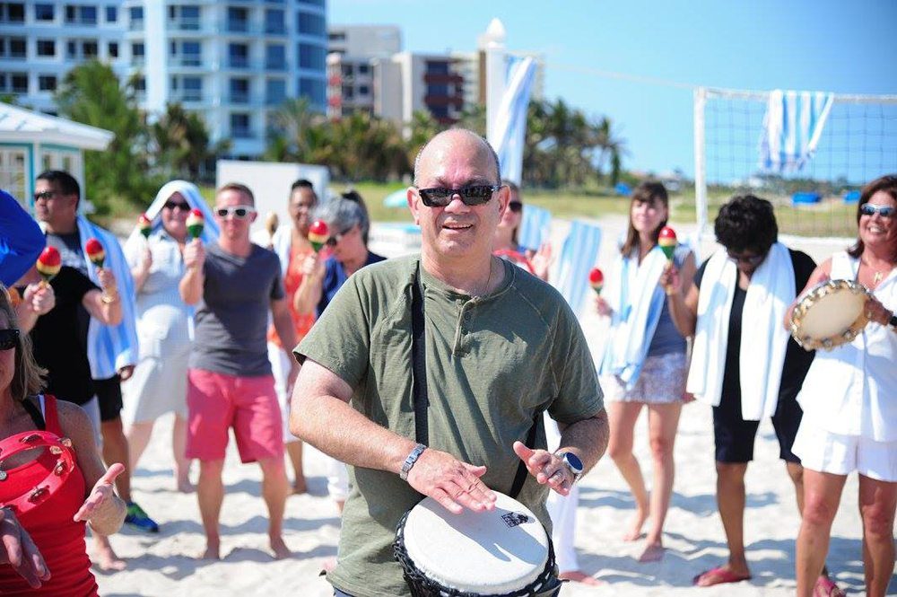 Participants engage in a lively Drumming Up A Team event on the beach, using percussion instruments to create a sense of unity and rhythm among the group in a sunny outdoor setting.