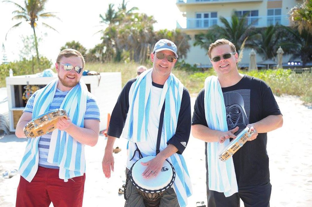 Three participants smiling and drumming together in a Drumming Up A Team event on the beach, wearing striped towels and enjoying the upbeat rhythm in the sunny outdoor setting.