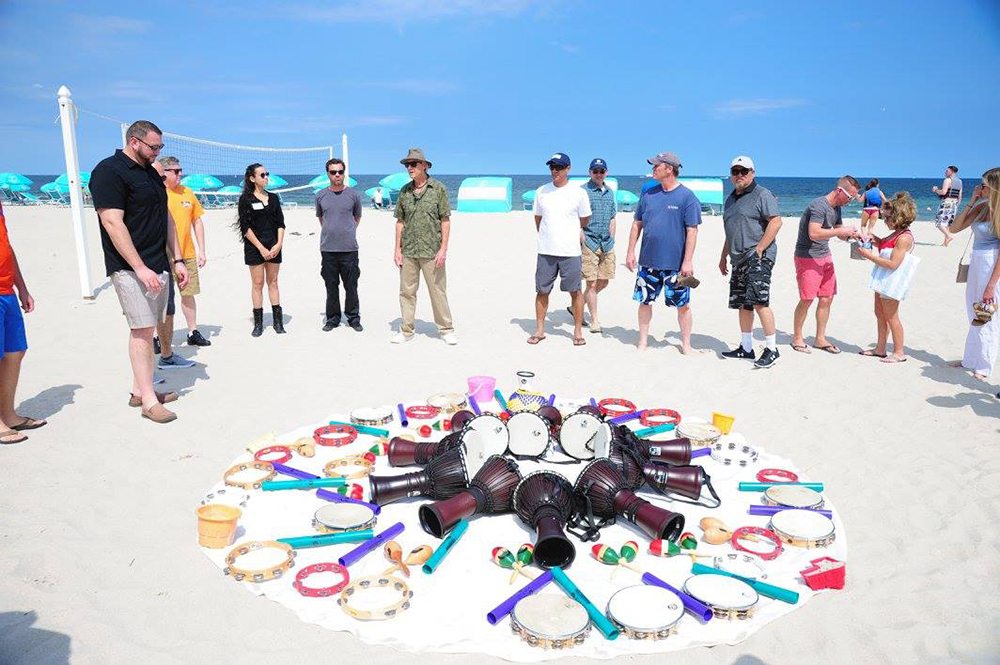 A vibrant array of percussion instruments arranged in a circle on the sand during a Drumming Up A Team event at the beach, with participants standing around preparing to play.