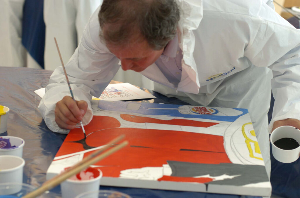 A participant wearing a white protective suit carefully paints a section of a mural during The Big Picture team building event. The individual focuses on adding details to a vibrant red, blue, and white canvas, contributing to the collaborative masterpiece.