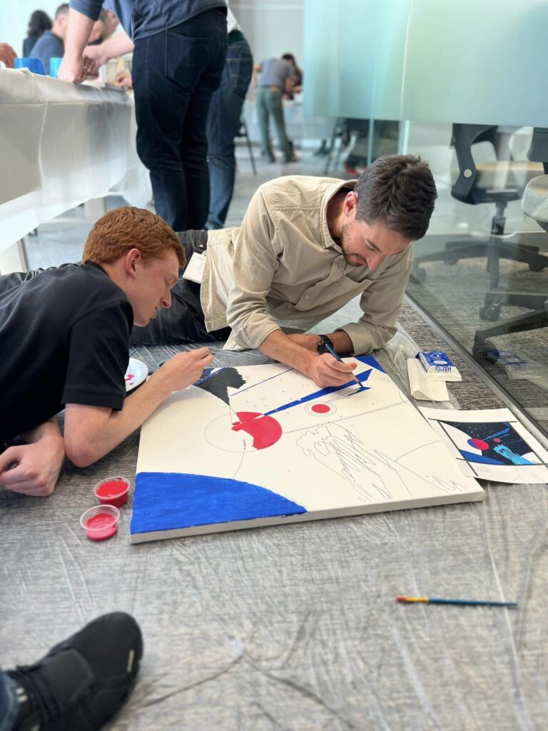 Two participants are lying on the floor, working together to paint a section of the canvas during The Big Picture team building event. They focus intently on adding details and colors, collaborating on their contribution to the larger mural.