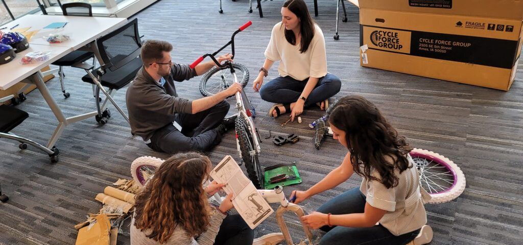 A group of four people sitting on the floor assembling a bicycle during a Charity Bike Build event. They are carefully reading instructions and working collaboratively, with bike parts and tools spread around them.