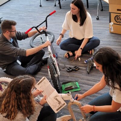 A group of four people sitting on the floor assembling a bicycle during a Charity Bike Build event. They are carefully reading instructions and working collaboratively, with bike parts and tools spread around them.