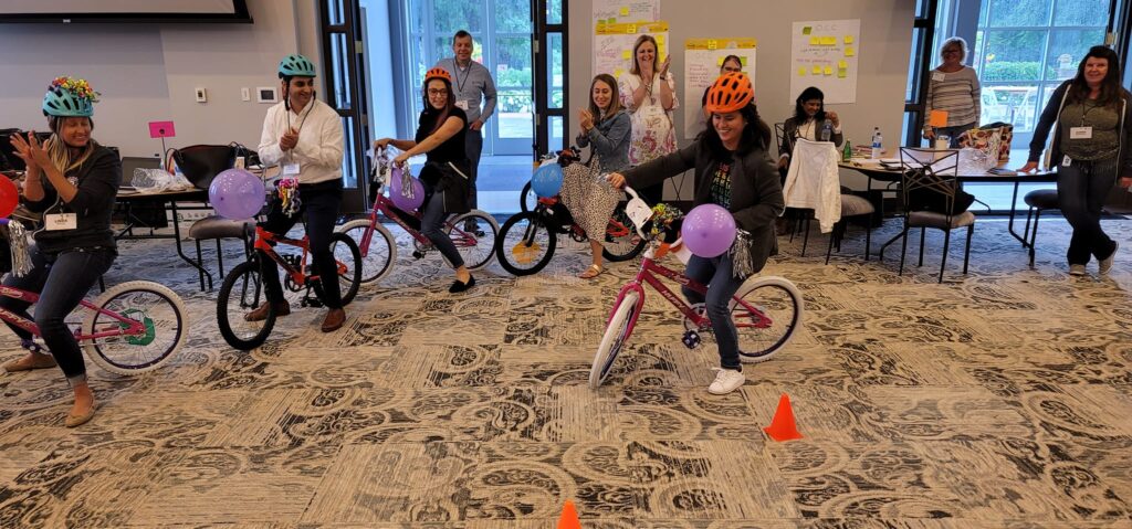 Participants wearing colorful helmets riding their newly assembled bikes during a Charity Bike Build event. The bikes are decorated with balloons and streamers as the participants test their creations, navigating through cones in a fun indoor obstacle course. Team members cheer and applaud from the background.