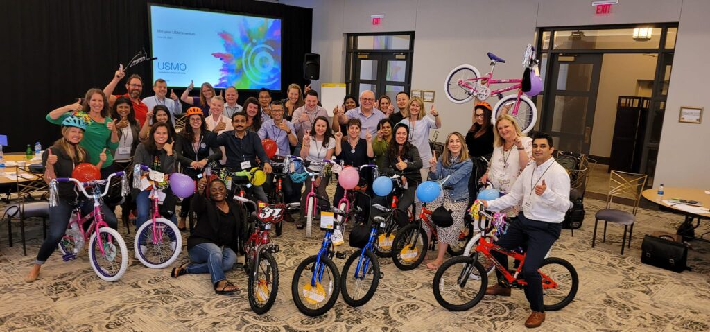 Large group of smiling participants gathered around a collection of brightly colored children's bicycles during a Charity Bike Build event. Many participants are holding up the bikes they built, decorated with balloons and streamers, showcasing the teamwork and joy of the event.