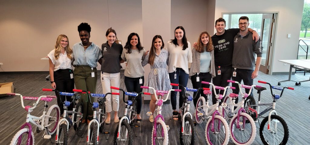 A group of smiling participants standing behind a row of assembled children's bicycles during a Charity Bike Build event. The bikes are pink and blue with training wheels, and the group appears happy with their completed team project.