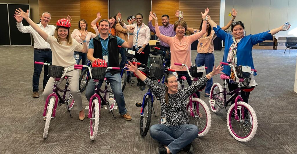 A group of excited participants proudly displaying their completed bicycles during a Charity Bike Build event. They stand behind and beside the assembled bikes with their arms raised in celebration.