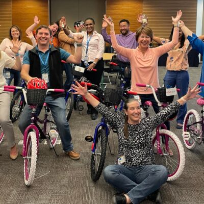 A group of excited participants proudly displaying their completed bicycles during a Charity Bike Build event. They stand behind and beside the assembled bikes with their arms raised in celebration.