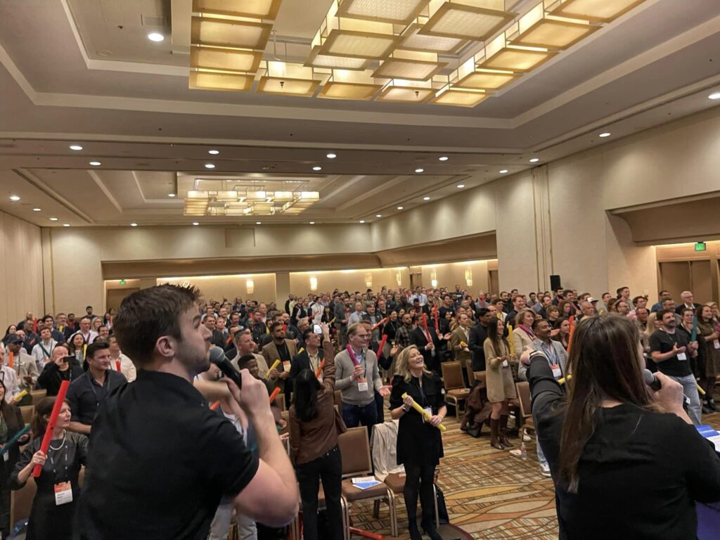 A large group participates in the Boom Time team building activity, guided by energetic facilitators. The crowd enthusiastically follows along with musical cues, using colorful Boomwhackers to create rhythmic sounds together.