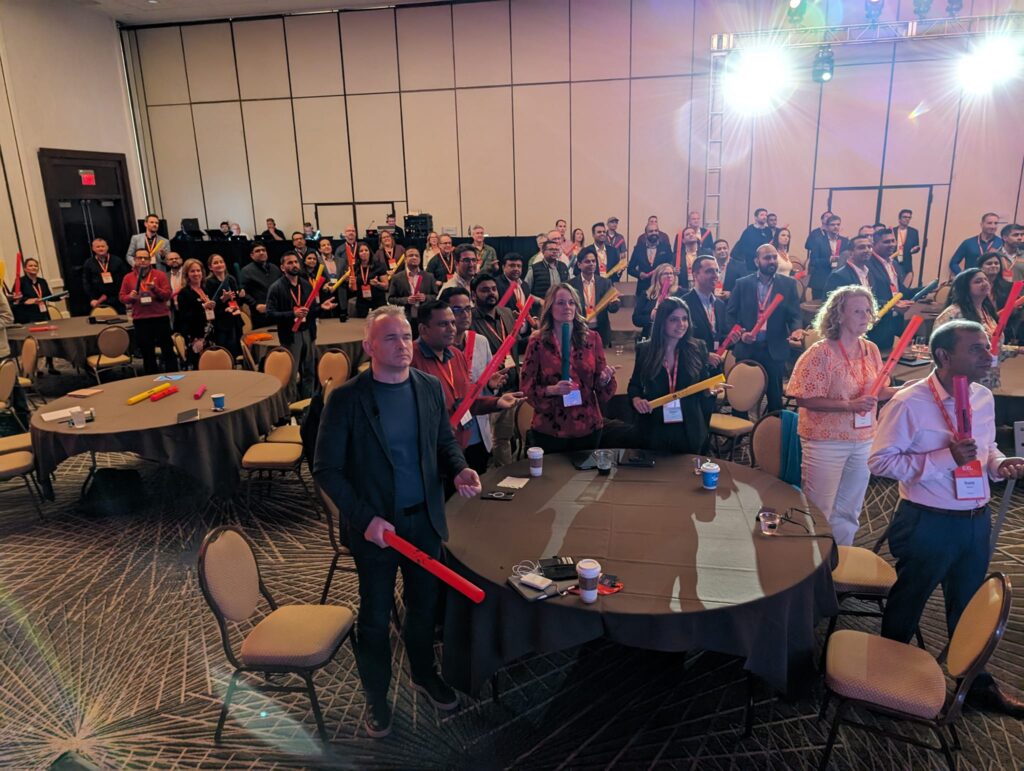 Conference attendees stand at their tables holding colorful Boomwhackers, engaged in a fun, interactive Boom Time team building session, focused on rhythm and collaboration.