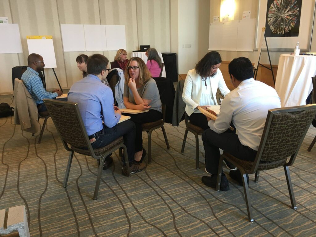 Small groups of participants engage in one-on-one discussions while holding wooden boards during a board-breaking team building activity focused on overcoming personal barriers.