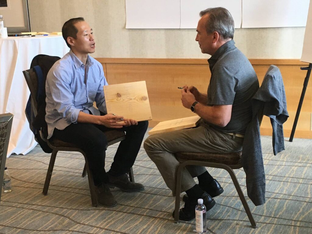 Two participants sit facing each other, discussing their wooden boards during a transformational team building experience centered on breaking through barriers.