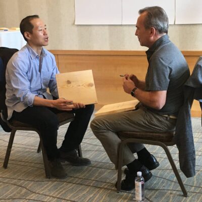 Two participants sit facing each other, discussing their wooden boards during a transformational team building experience centered on breaking through barriers.