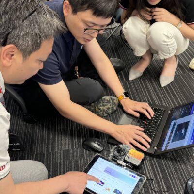 A team building activity in progress, with participants collaborating using laptops and tablets to create their AI-powered broadcasts.