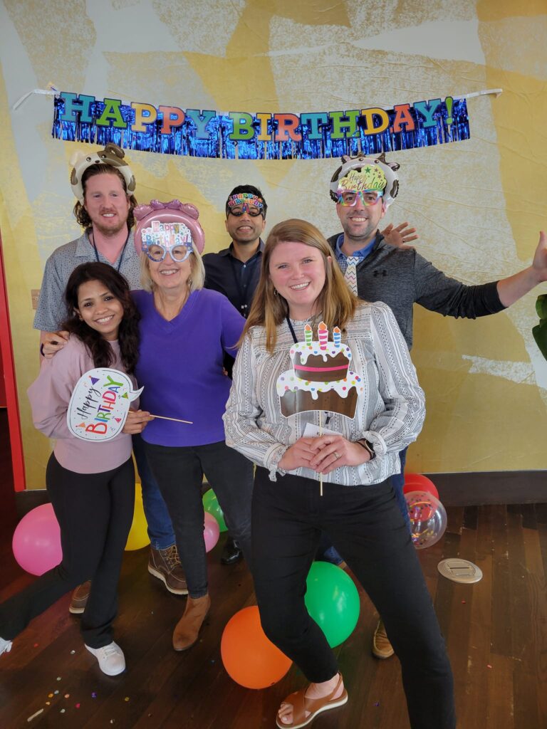 A group of six people poses happily in front of a 'Happy Birthday' banner. They're holding birthday-themed props, surrounded by colorful balloons, and wearing fun hats and party accessories during a team building event.