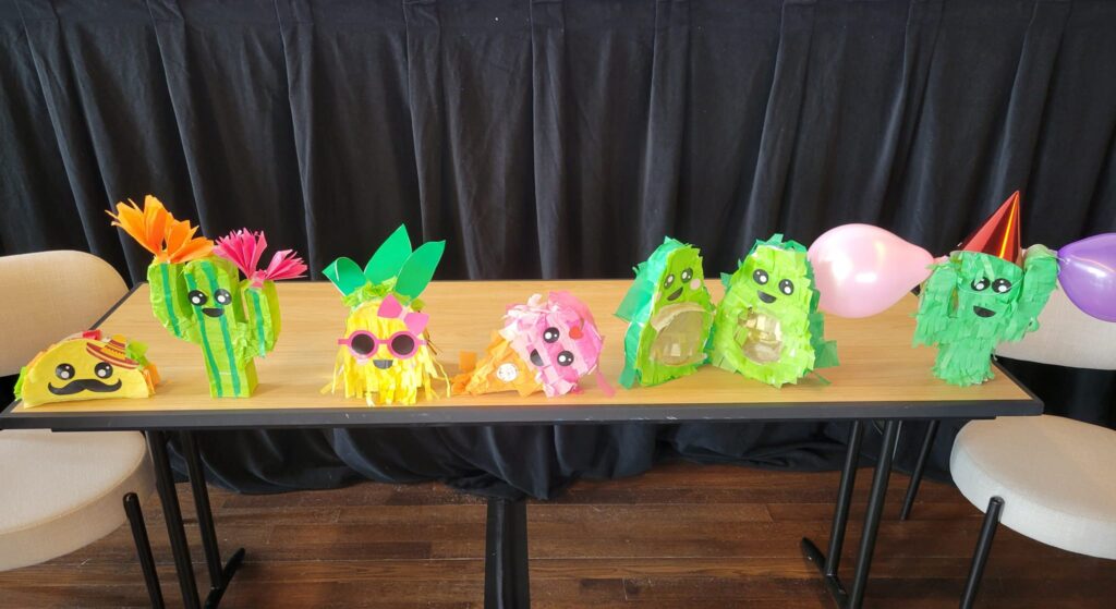 A row of colorful and playful piñatas, including cacti and other fun shapes, is displayed on a table. Some are wearing party hats, sunglasses, or decorated with balloons, bringing a festive vibe to the birthday kits that are going to be donated as a result of this team building activity.