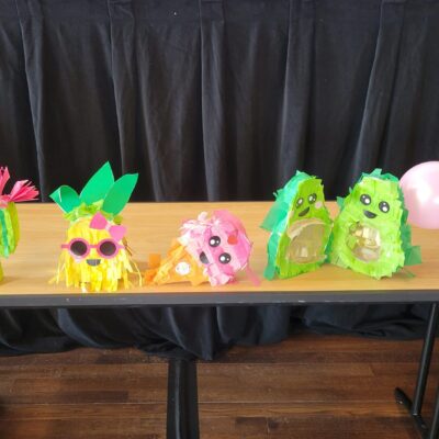 A row of colorful and playful piñatas, including cacti and other fun shapes, is displayed on a table. Some are wearing party hats, sunglasses, or decorated with balloons, bringing a festive vibe to the birthday kits that are going to be donated as a result of this team building activity.