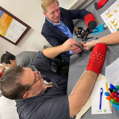 Two participants are actively assembling a prosthetic hand during The Prosthetic Hand Project. They are wearing red prosthetic simulation sleeves, adding a level of challenge and empathy to the task. One person is holding and connecting the components of the mechanical hand while the other assists. A variety of tools and assembly instructions are visible on the table, and a notebook and colorful pens are seen nearby, indicating careful planning and attention to detail. This image highlights the teamwork, focus, and effort involved in assembling prosthetic hands for donation to those in need.