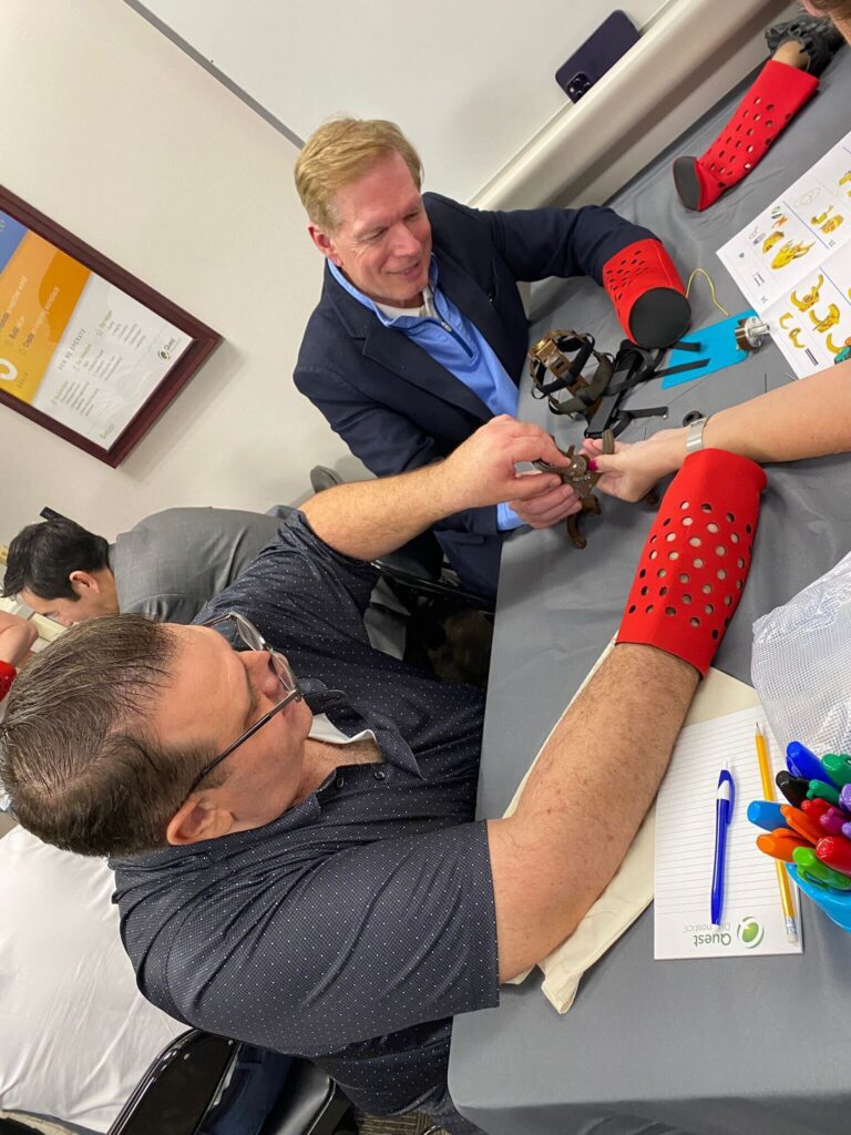 Two participants are actively assembling a prosthetic hand during The Prosthetic Hand Project. They are wearing red prosthetic simulation sleeves, adding a level of challenge and empathy to the task. One person is holding and connecting the components of the mechanical hand while the other assists. A variety of tools and assembly instructions are visible on the table, and a notebook and colorful pens are seen nearby, indicating careful planning and attention to detail. This image highlights the teamwork, focus, and effort involved in assembling prosthetic hands for donation to those in need.