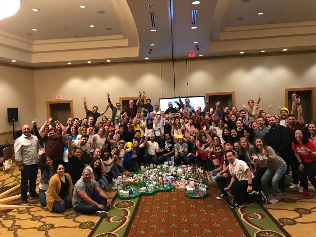 Large group celebrating their completed City Build project during a team building event, with a colorful miniature city displayed in the center of the room.