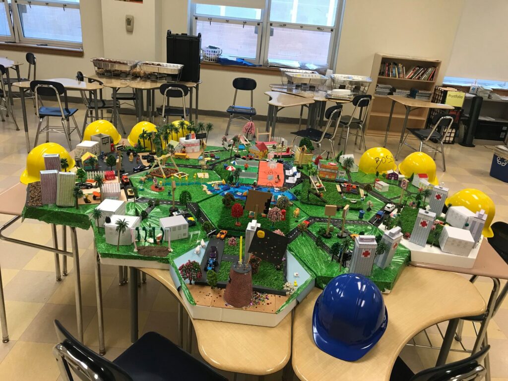 Colorful miniature city model displayed on tables, surrounded by yellow and blue hard hats, featuring buildings, roads, and greenery for a City Build team building activity.