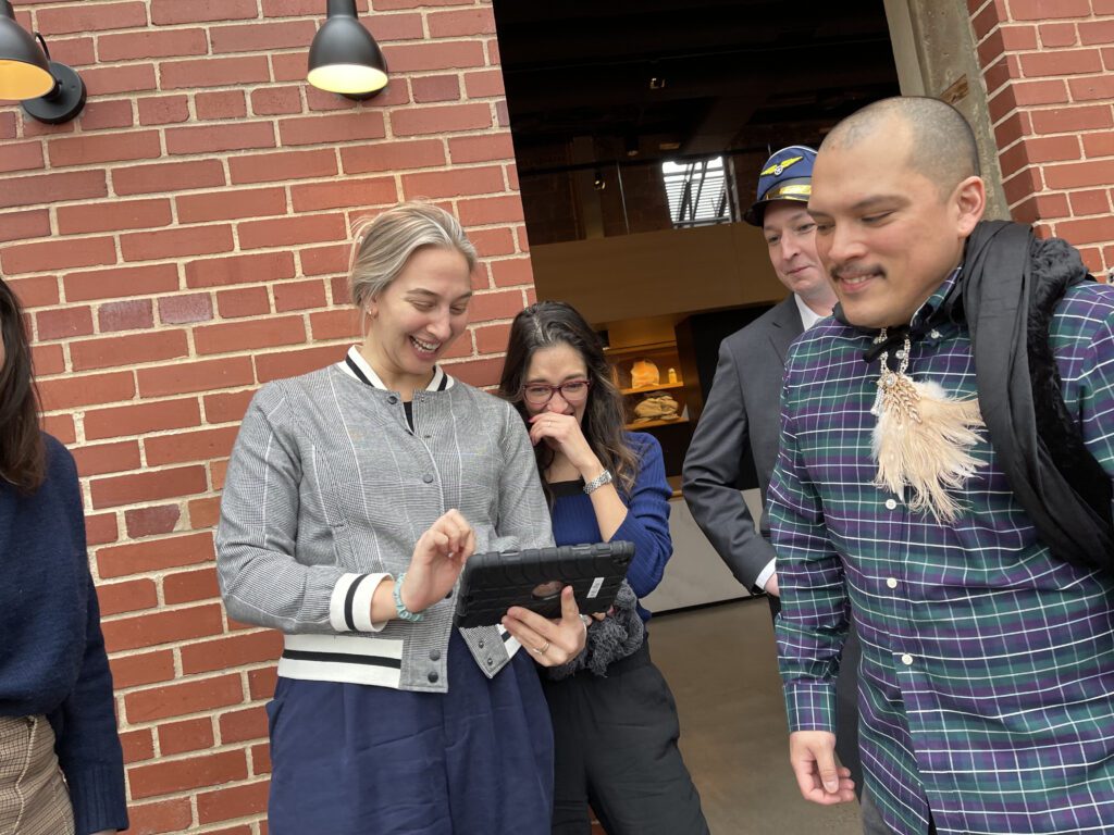 A group of colleagues smiling and interacting with a tablet during an outdoor team building exercise, demonstrating collaboration and teamwork while enjoying the making a movie activity.