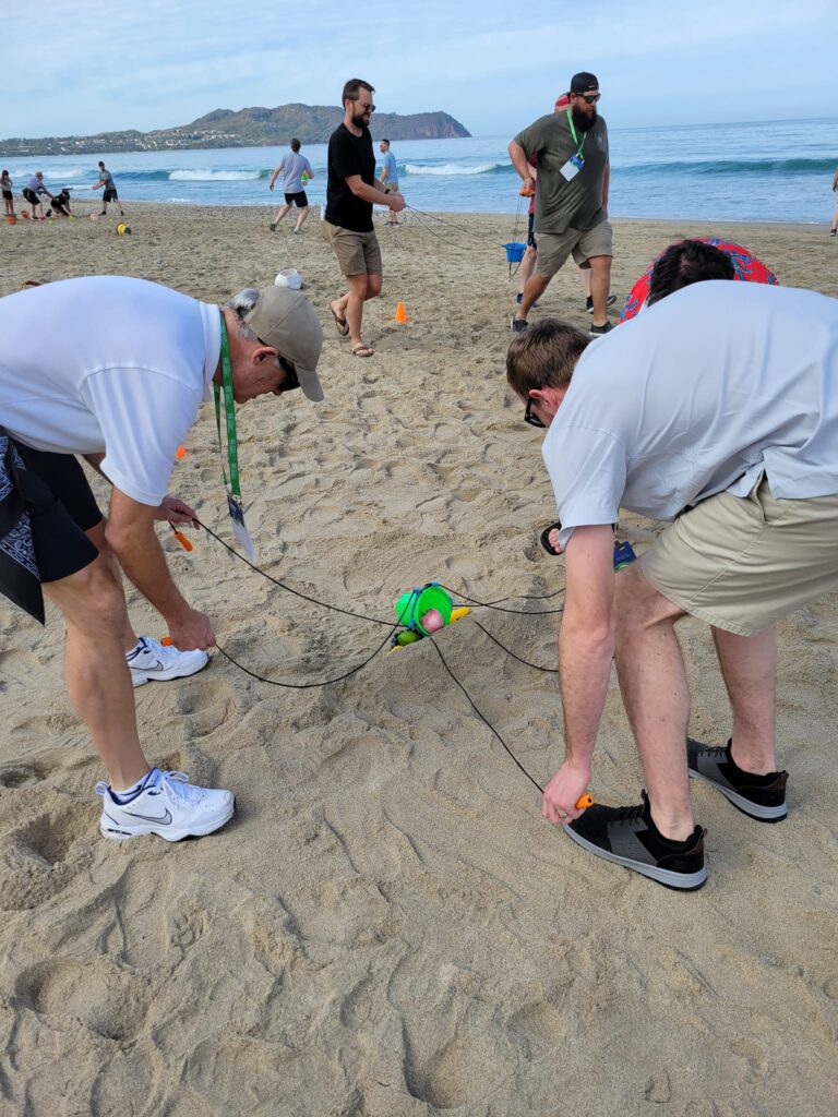 Two teammates work together using ropes to control a ball during a beach team building challenge.