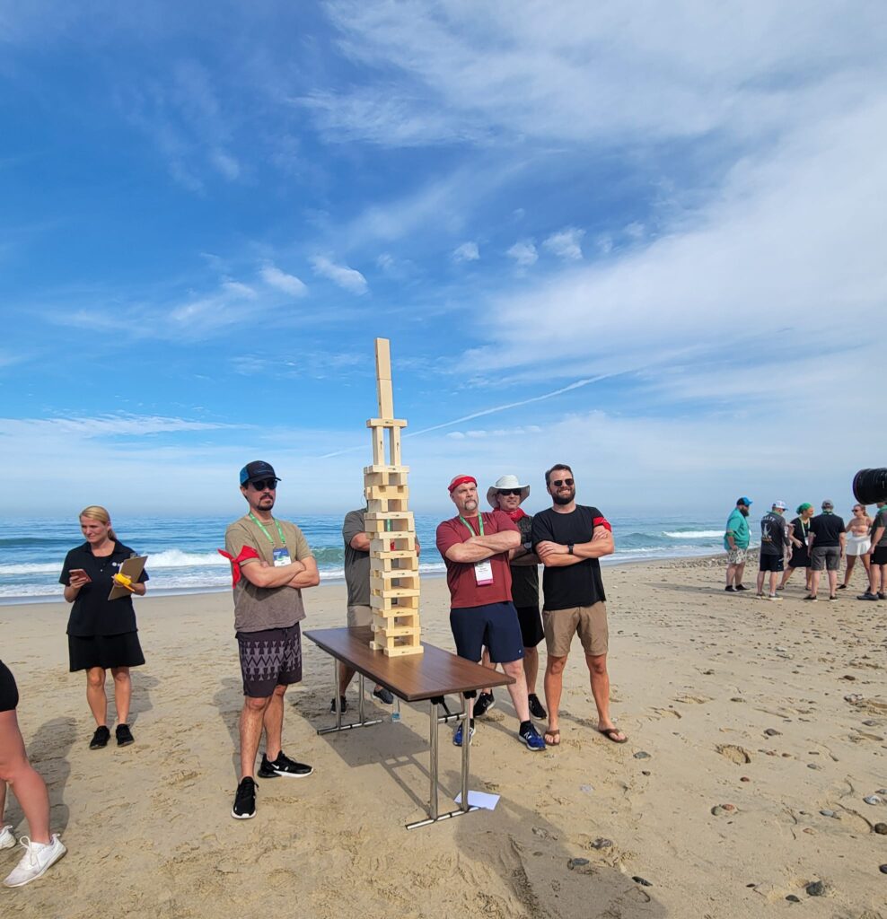 Team proudly stands next to their completed giant Jenga tower on the beach during a team building event.