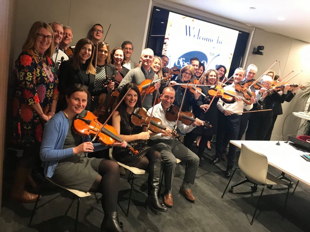 Group of participants playing violins together in a meeting room during the Crescendo team building event, smiling and enjoying the collaborative musical activity.