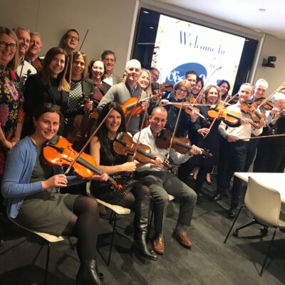 Group of participants playing violins together in a meeting room during the Crescendo team building event, smiling and enjoying the collaborative musical activity.