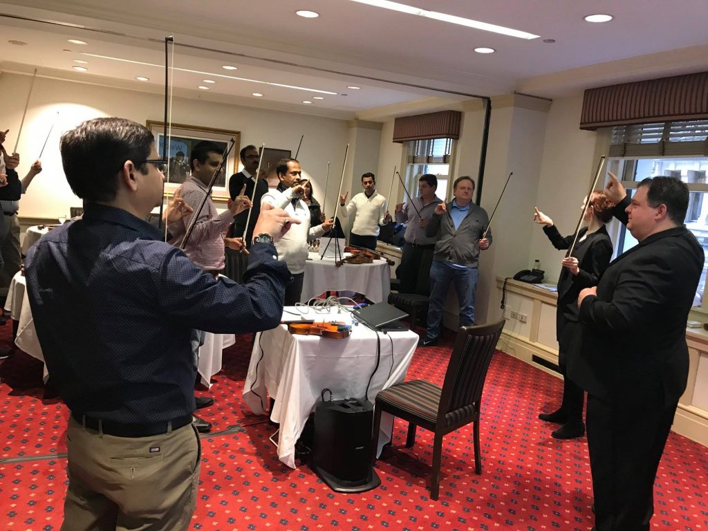 Participants in a Crescendo team building event stand in a circle, holding violins and bows, as an instructor leads them through a collaborative music lesson in a conference room.