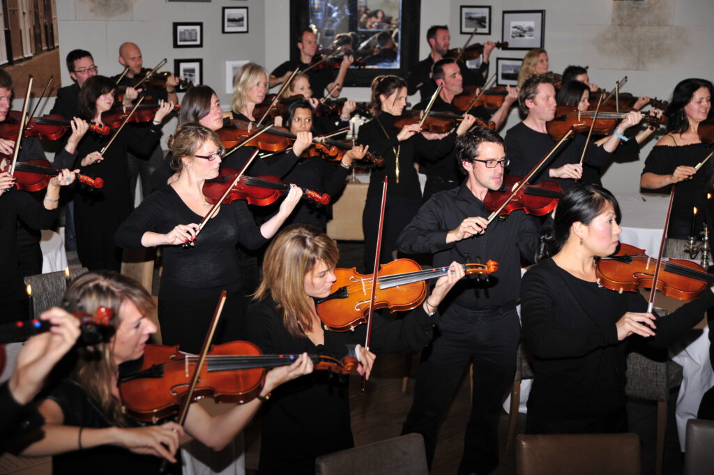 Participants in black clothing perform together as a string orchestra during a Crescendo team building event, holding violins and violas with focused expressions.