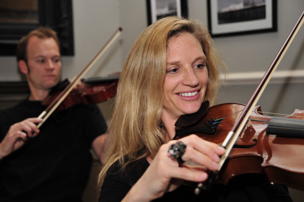 Smiling participants learning to play violins together during the Crescendo team building event, highlighting teamwork and musical collaboration.