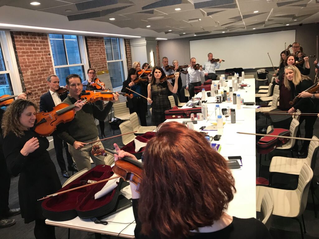 Participants in a Crescendo team building event practicing their violins together in a bright, modern conference room, guided by instructors.